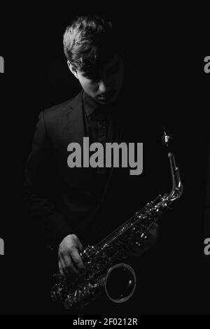 Un colpo della scala di grigi di un ragazzo freddo e bello che tiene il suo sassofono su sfondo scuro Foto Stock