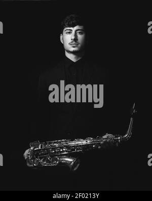 Un colpo della scala di grigi di un ragazzo freddo e bello che tiene il suo sassofono su sfondo scuro Foto Stock