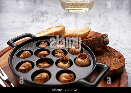 Lumache fritte, escargotnière in padella, con crostini, su tavola di legno, su fondo chiaro, con un bicchiere di vino bianco Foto Stock