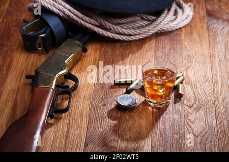 Old West fucile e munizioni con bicchiere di whisky e. ghiaccio con vecchie monete di dollari d'argento su tavolo di legno da bar Foto Stock