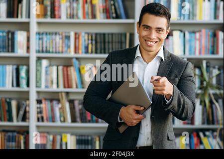 Un imprenditore di stile soddisfatto sta in piedi in ufficio, in posa con ampio sorriso. Bell'uomo d'affari ispanico che guarda direttamente la fotocamera, tenendo il computer portatile in braccio Foto Stock