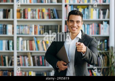 Ritratto di gioioso dipendente o studente di stile maschile in una biblioteca. Felice uomo d'affari ispanico in abito elegante sorridente, guardando direttamente la fotocamera, in posa in ufficio moderno Foto Stock