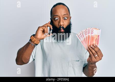 Giovane afroamericano che parla sullo smartphone tenere in mano banconote norvegesi di corona facendo il pesce faccia con la bocca e. squinning e Foto Stock