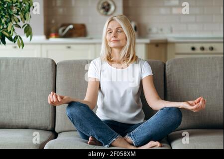 Calma attraente donna di mezza età si siede sul divano a casa e freddo. La donna anziana è pratica yoga e meditazione in posizione di loto con gli occhi chiusi, alleviare lo stress, concetto di calma Foto Stock