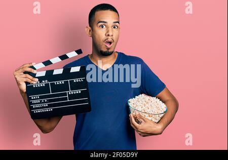 Giovane afroamericano mangiare popcorn tenere clapboard film paura e scioccato con sorpresa e stupito espressione, paura e volto eccitato. Foto Stock