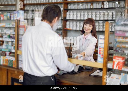 Felice sicuro giovane donna farmacista in piedi dietro il bancone e offrendo la scatola della medicina, parlando con il cliente maschio anziano. Vendita di medicina Foto Stock