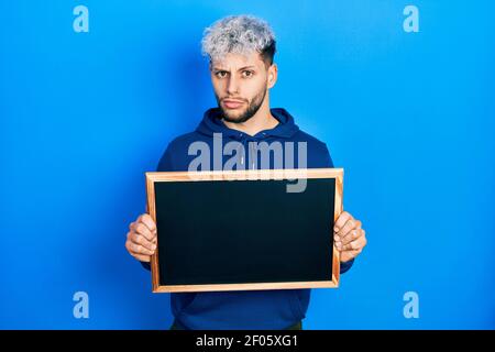 Giovane uomo ispanico con capelli tinti moderni che tengono nero tavola indistinto e confuso espressione. Concetto di dubbio. Foto Stock