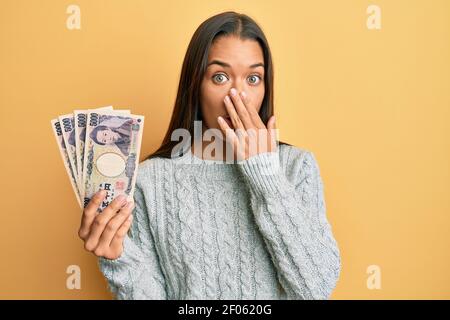 Bella donna ispanica che tiene 5000 yen giapponesi banconote che coprono la bocca con la mano, scioccata e impaurita per errore. Sorpresa espressione Foto Stock