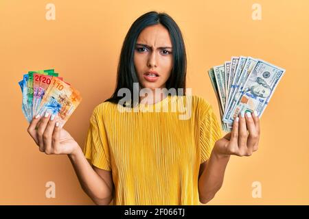 Giovane bruna donna che tiene un mazzo di dollari e franchi svizzeri banconote in shock viso, guardando scettico e sarcastico, sorpreso con bocca aperta Foto Stock