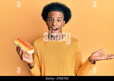 L'uomo afroamericano con i capelli afro che mangiano hotdog festeggia il successo con sorriso felice e espressione vincente con mano sollevata Foto Stock