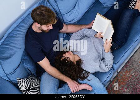 Da sopra giovane donna in vestiario casual sdraiato sul divano e libro di lettura mettendo la testa sulle ginocchia del ragazzo Foto Stock