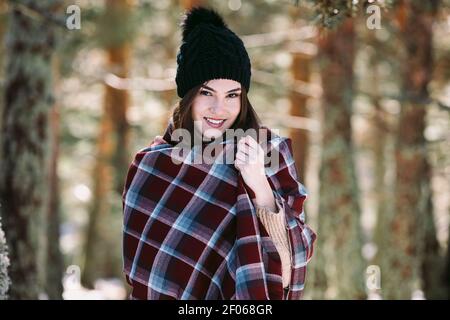 Contenuto femmina avvolto in caldo plaid in piedi in boschi innevati e guardando la fotocamera Foto Stock
