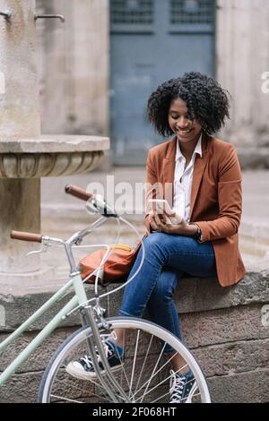 Giovane donna afroamericana in elegante abito casual di navigazione mobile telefono mentre si siede vicino alla bicicletta sul vecchio bordo della fontana di pietra in città Foto Stock