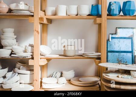 Raccolta di ciotole e vasi in ceramica fatti a mano con pentole e. piastre con vecchio specchio vicino a diversi tipi di utensili in piedi su scaffali di legno in ligh Foto Stock