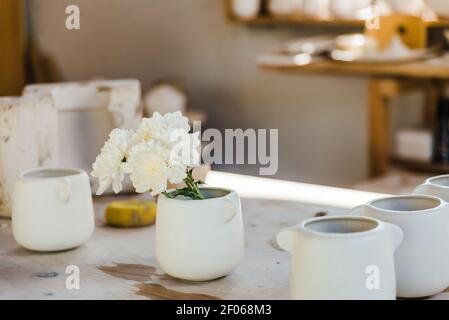 Set di tazze in ceramica bianca con fiori posti sul bancone in studio luminoso vicino ad oggetti per artigianato Foto Stock