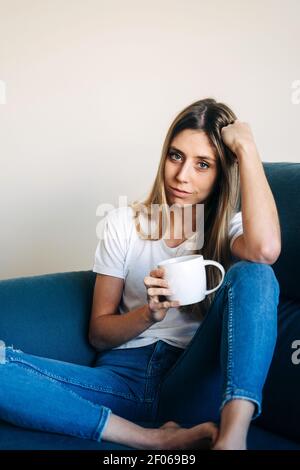 Donna pensiva con tazza di caffè guardando la macchina fotografica e. appoggiandosi sulla mano mentre si siede sul divano con la gamba piegata Foto Stock