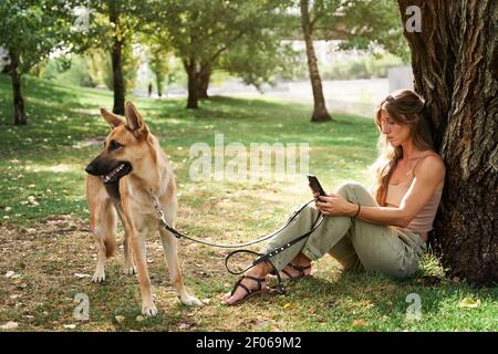 Donna con capelli lunghi seduta sull'erba vicino al Pastore tedesco animali domestici e uso del telefono cellulare nel parcheggio Foto Stock