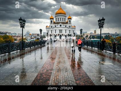 Persone che camminano sotto la pioggia sul Ponte Patriarshiy sopra il Fiume Moskva verso la Cattedrale di San Salvatore con il cielo tempestoso. Mosca, Russia Foto Stock