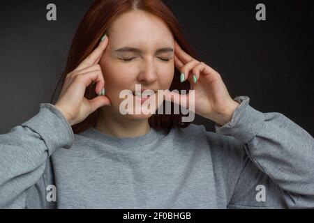 Giovane ragazza dai capelli rossi su sfondo nero in una T-shirt grigia, spreme templi, testa fa male Foto Stock