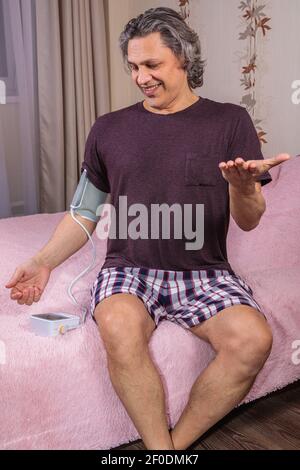 Un uomo di 50 anni misura la sua pressione sanguigna con un monitor della pressione sanguigna a casa, seduto sul letto nei vestiti di casa. Sorpreso dallo strumento Foto Stock