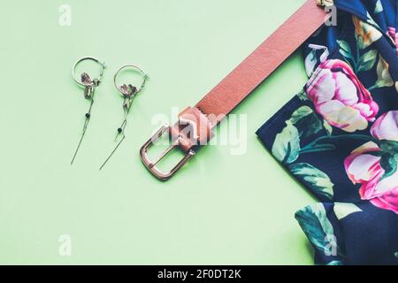 Vista dall'alto degli accessori estivi per la donna moderna in vacanza. Abito colorato, cintura in pelle marrone e orecchini in oro su sfondo verde. Spazio di copia. Foto Stock