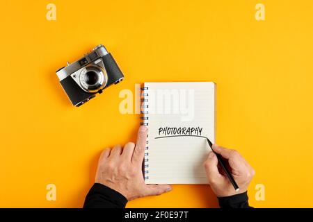 Mano maschile scrivere la parola fotografia in un notebook accanto a una vecchia fotocamera vintage su sfondo giallo. Foto Stock