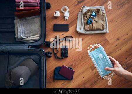 Il viaggiatore si sta preparando per viaggiare nella nuova normalità. Maschere per il viso e igienizzatore per le mani per una protezione personale contro l'imballaggio di suitcace con sume Foto Stock