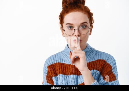 Shhh il suo un segreto. Carino ragazza hipster rosso shushing, chiedendo di essere tranquilla o rimanere in silenzio, facendo gesto tabù, in piedi contro sfondo bianco Foto Stock