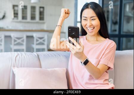 La donna asiatica molto gioiosa tiene uno smartphone seduto sul divano a casa, guarda lo schermo e celebra il buon affare o le notizie, alzata mano estatica, vittoria e concetto trionfo Foto Stock