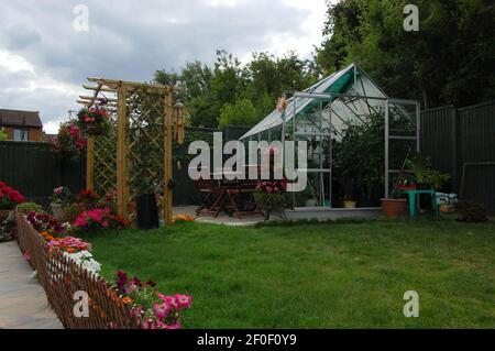 Serra in giardino erba percorso pavimentazione pietre recinto scherma albero alberi vaso piante pentole porta aperta cestini appesi colore cielo pioggia Foto Stock