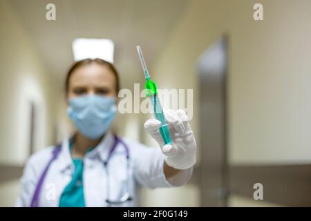 Una dottoressa in una maschera protettiva sul corridoio dell'ospedale. Nelle sue mani tiene una siringa con un vaccino incandescente. Mettere a fuoco la siringa. C Foto Stock