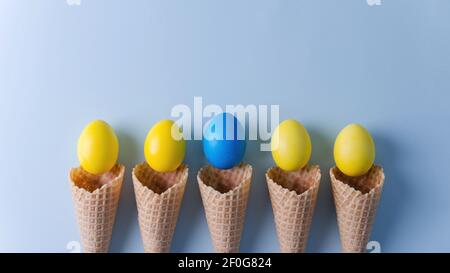 Le uova gialle e una blu in coni di waffle giacciono su uno sfondo blu orizzontale. Sfondo pasquale concetto piatto, copia spazio, fuoco selettivo Foto Stock