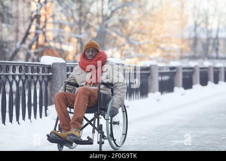 Ritratto a lunghezza intera dell'uomo afroamericano che usa la sedia a rotelle all'aperto in inverno e guardando la macchina fotografica, spazio di copia Foto Stock