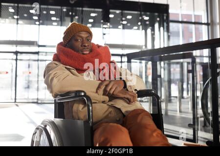 Ritratto dell'uomo afro-americano in sedia a rotelle guardando la macchina fotografica nel centro commerciale accessibile o nella stazione urbana della città, copy space Foto Stock