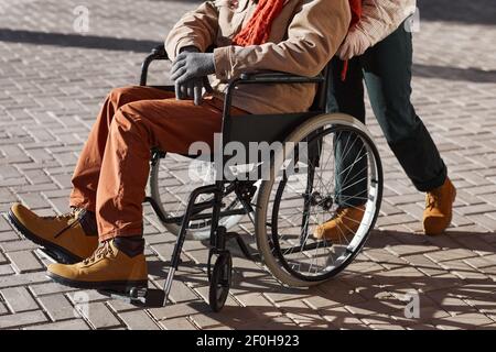 Ripresa di un irriconoscibile uomo afroamericano in sedia a rotelle che si muove in città urbana illuminata dalla luce del sole con la donna che assiste, copia spazio Foto Stock