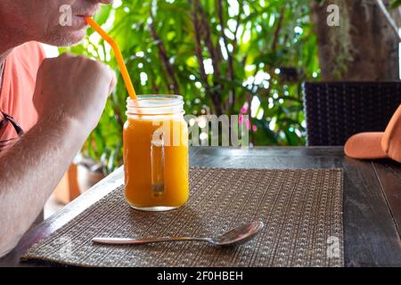 L'uomo adulto beve un frullato di mango attraverso una paglia, una deliziosa bevanda fredda alla frutta in un caffè in Thailandia, vacanza estiva Foto Stock