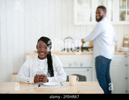 Giovane donna nera seduta in cucina con smartphone mentre marito Pranzo di cucina Foto Stock