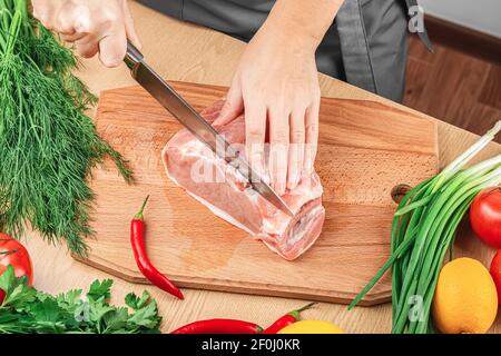 Le mani femminili tengono un coltello in mano e tagliano un pezzo di carne di maiale fresca su una tavola di legno sullo sfondo di erbe e verdure fresche. Vista dall'alto, c. Foto Stock