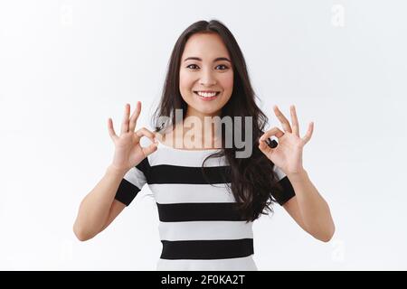 Ragazza che prende tutto sotto controllo, nessuna preoccupazione. Affascinante giovane donna asiatica in t-shirt a righe vi assicura, incoraggiano evento è andato grande, mostrando ok Foto Stock