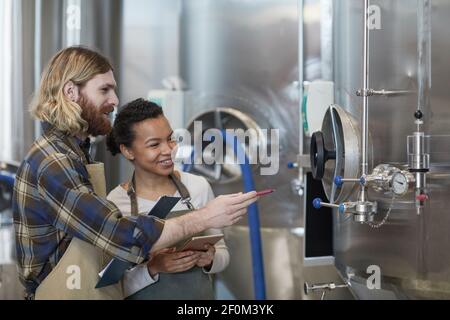 Vista laterale ritratto di due giovani lavoratori sorridenti che ispezionano la produzione presso la moderna fabbrica di birra artigianale, spazio copia Foto Stock
