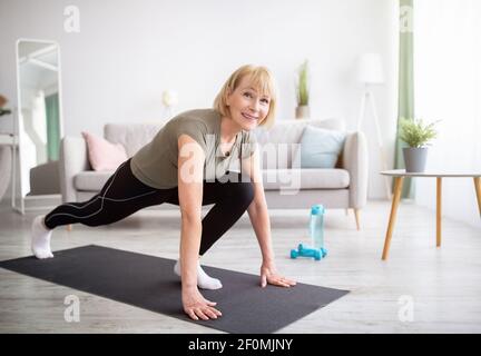 Donna anziana atletica positiva che fa la posa yoga affondo del corridore sull'allenamento domestico, spazio di copia Foto Stock