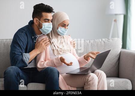 Interessato coppia musulmana incinta in maschere mediche usando il laptop a. Casa Foto Stock
