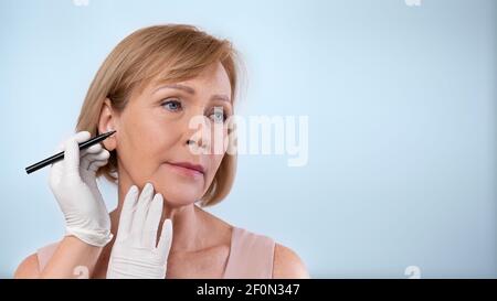 Chirurgo di plastica che disegna linee sul viso della donna matura per l'operazione cosmetica su sfondo blu studio, banner design Foto Stock