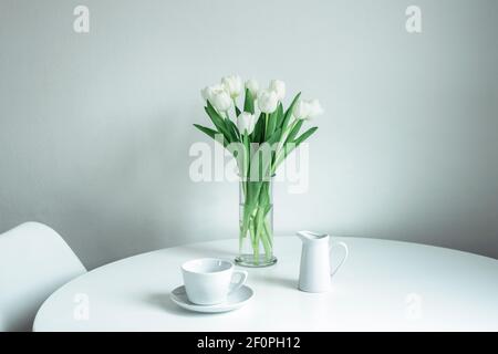 Mazzo di tulipani bianchi in un vaso di vetro, una tazza e una caraffa di latte su un tavolo bianco. Mattina, colazione, concetto di primavera. Foto Stock