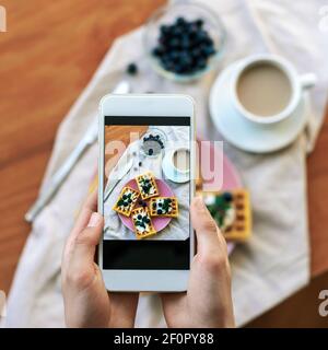Le mani della ragazza scattano foto di cialde salutari con mirtilli per telefono, vista dall'alto. Foto Stock