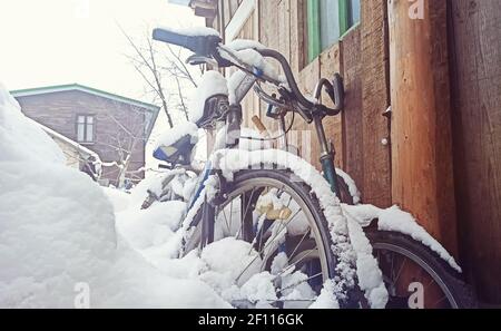 Biciclette coperte di neve si erigono contro un muro di legno vicino alla casa. Vecchie biciclette abbandonate nel cortile di una casa sotto la neve Foto Stock