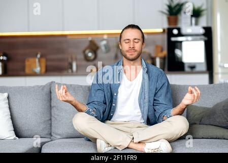 Calmo e spensierato ragazzo caucasico vestito in un elegante abbigliamento casual medita mentre si siede in posizione lotus sul divano a casa con gli occhi chiusi, rilassante. Concetto di calma e armonia Foto Stock