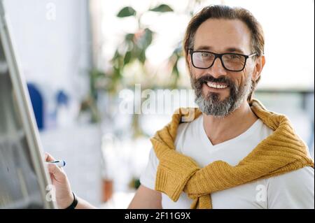Ritratto di un bel riuscito caucasico di mezza età. Insegnante con bearded vestito elegante o tutor di affari con gli occhiali scrive sulla lavagna a fogli mobili, sorride amichevole e guarda direttamente nella macchina fotografica Foto Stock