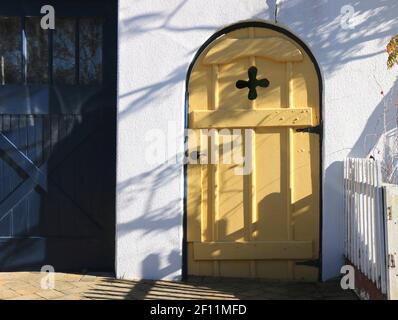 Bella vecchia porta di legno, gialla, arrotondata con una croce insettata, posta in una parete bianca di stucco, Monterey, CA. Foto Stock