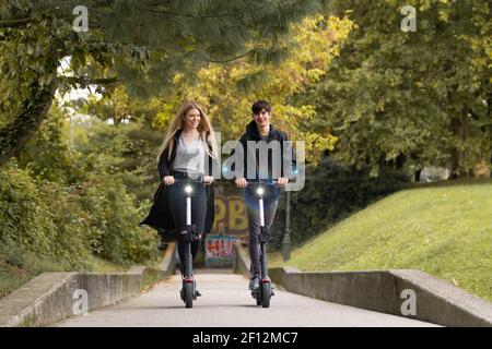 Adolescenti fashinable alla moda che cavalcano scooter elettrici a noleggio pubblici nel parco urbano della città. Nuovo trasporto pubblico ecologico e moderno a Lubiana Foto Stock